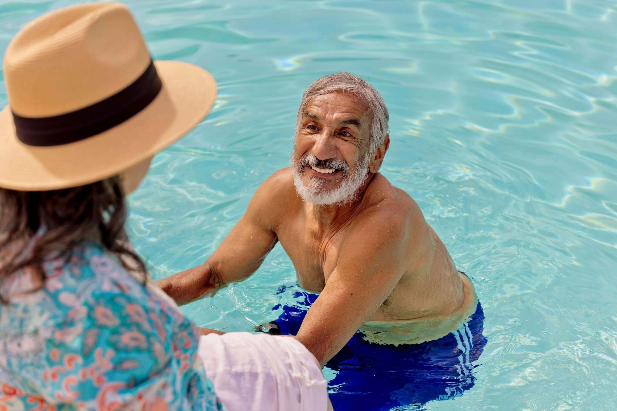 Senior man in the pool smiling at a senior woman wearing a sun hat