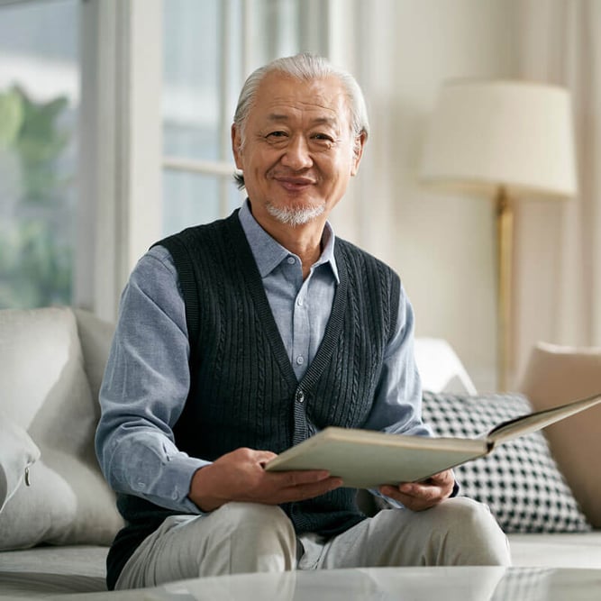 senior man sitting on couch holding a book and smiling at the camera